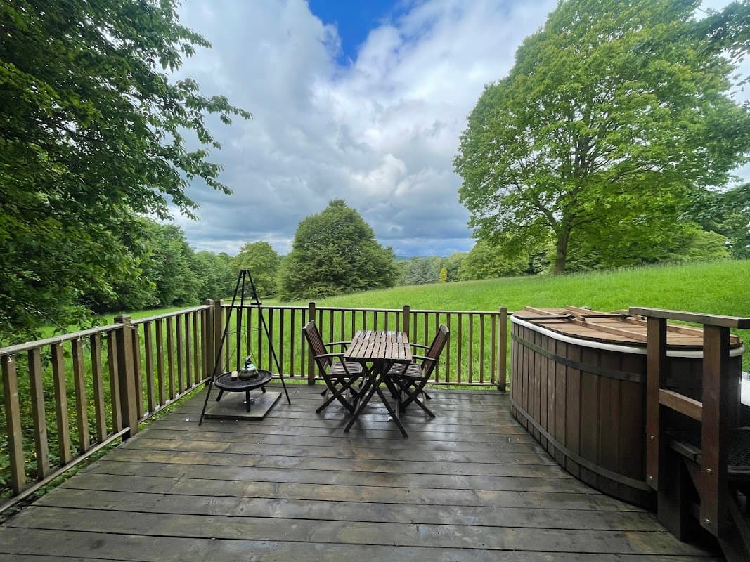 Wooden deck with a table and chairs, surrounded by lush greenery and a hot tub under a cloudy sky.