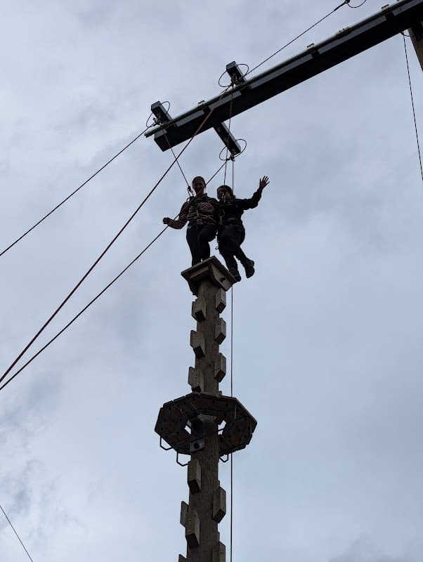 Two people standing atop a tall wooden climbing structure against a cloudy sky, with ropes and pulleys visible.