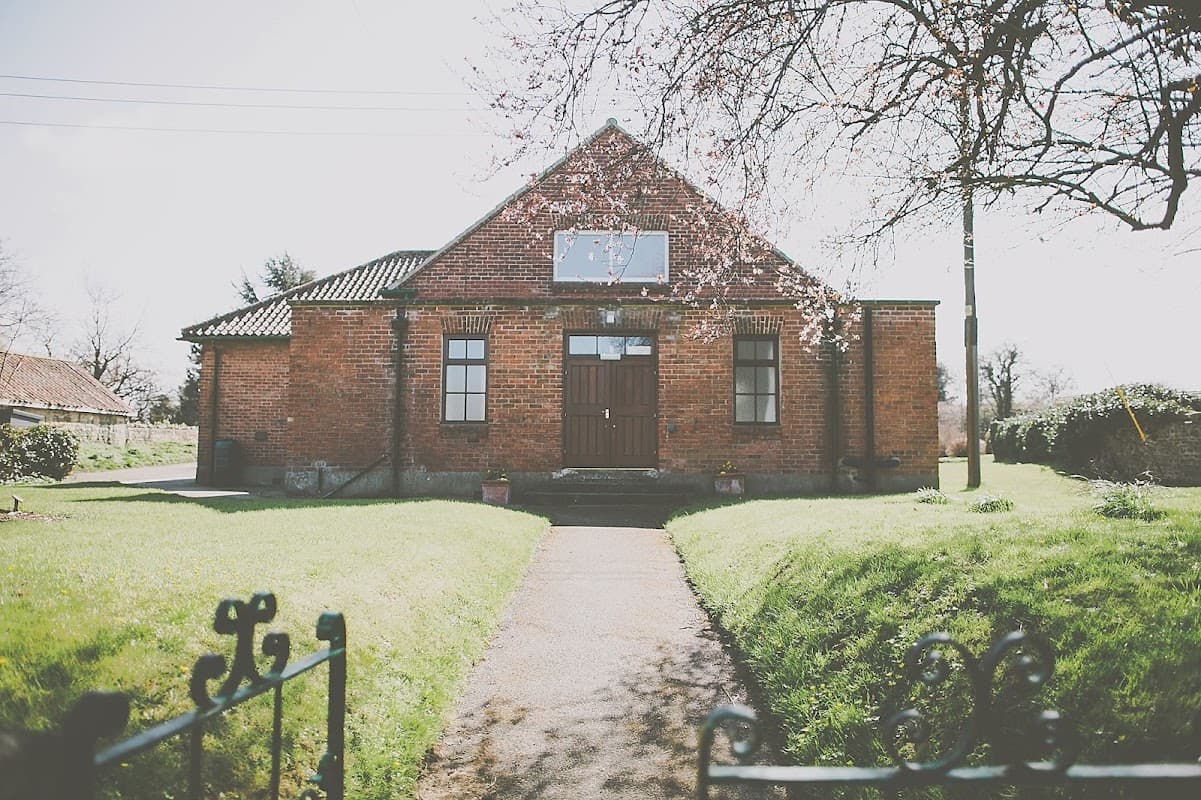 Red brick village hall with a central wooden door, surrounded by green grass and trees under a clear sky.