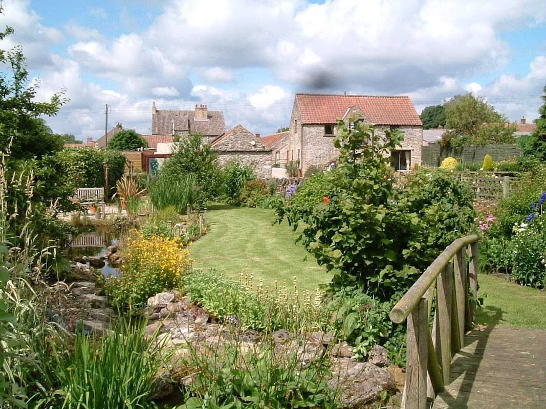 Lush garden with flowers, stone cottages, and a wooden bridge under a cloudy sky in Newton-upon-Rawcliffe, Yorkshire.