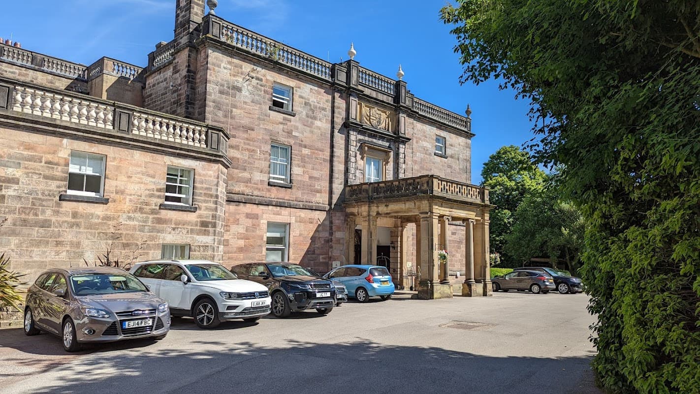 Historic stone building with ornate entrance, surrounded by greenery and parked cars in a sunny setting.