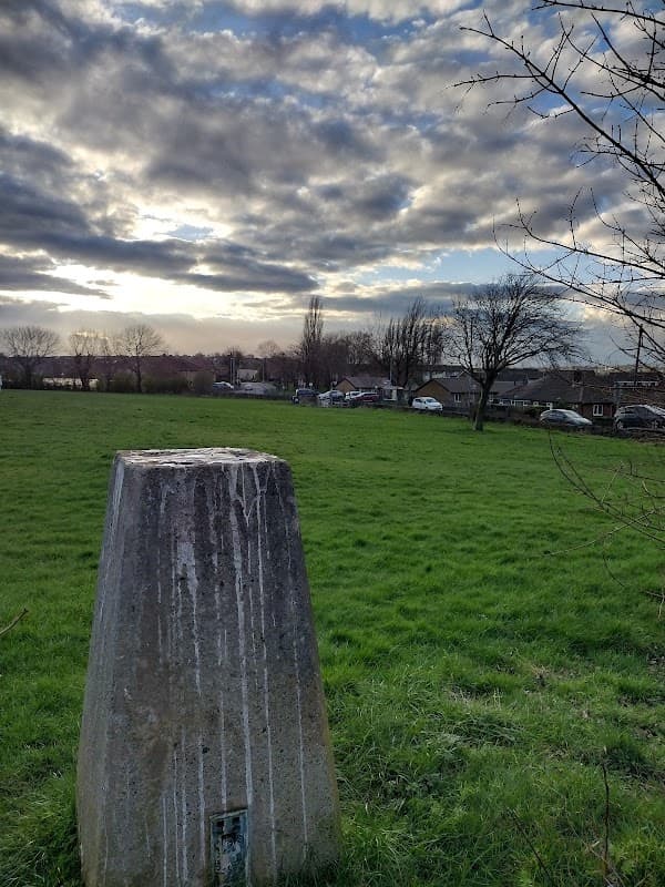 Concrete marker in grassy field under a cloudy sky, with trees and houses visible in the background.
