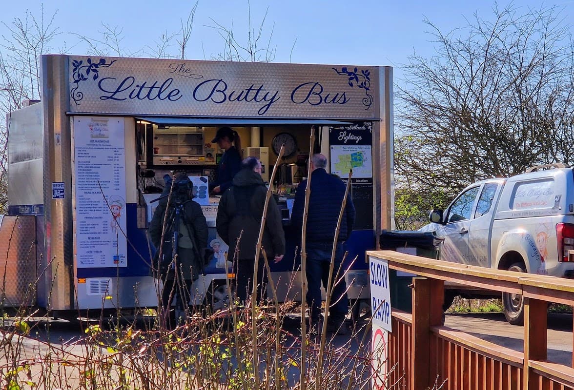 A food truck named "The Little Butty Bus" with customers ordering, surrounded by trees and a parked vehicle.