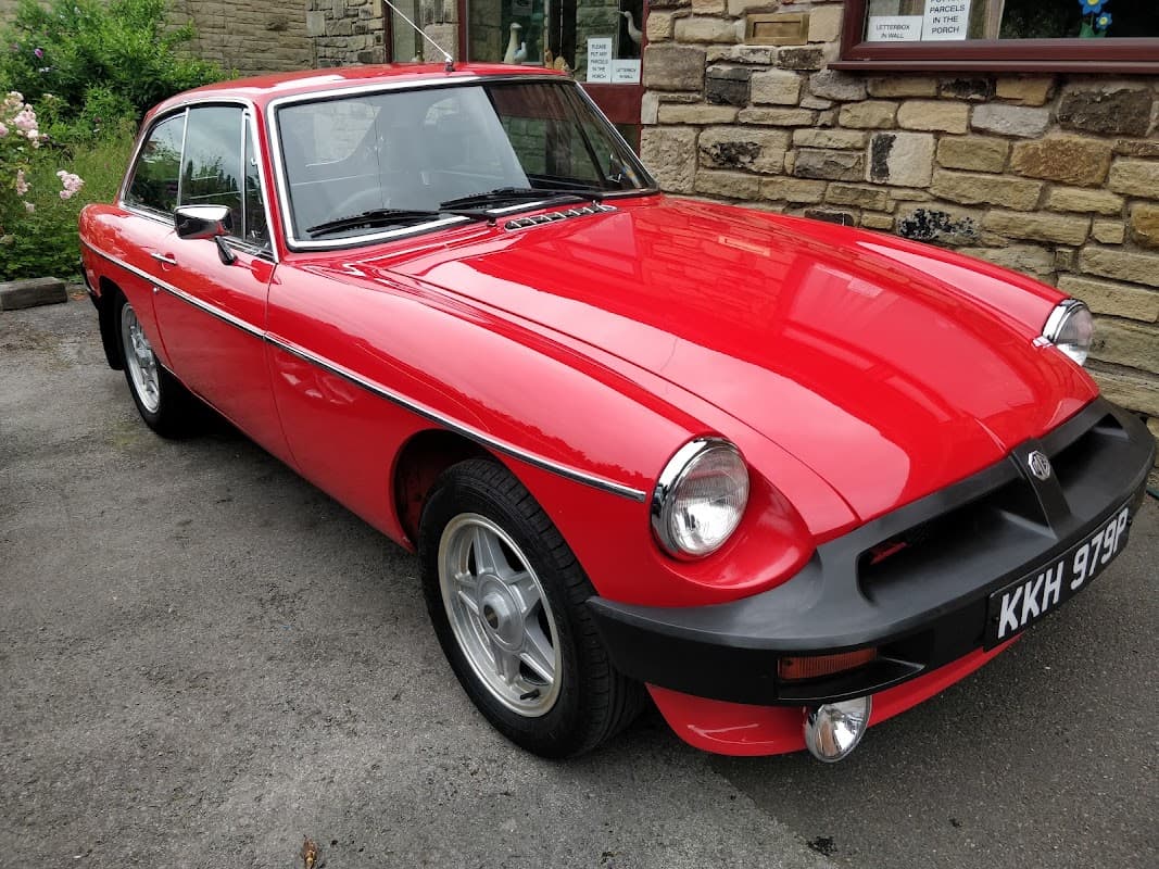 Red classic car parked in front of a stone building with windows, surrounded by greenery.