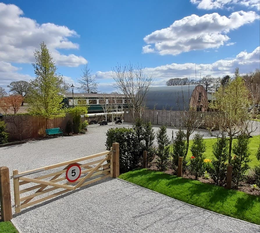 Gravel pathway leading to a wooden gate numbered 5, surrounded by greenery and a modern building under a blue sky.