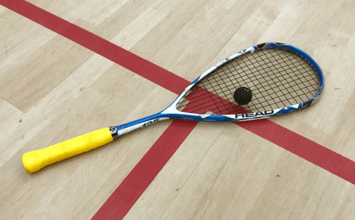 A squash racket with a yellow grip lies on a wooden court beside a black squash ball, marked by red lines.