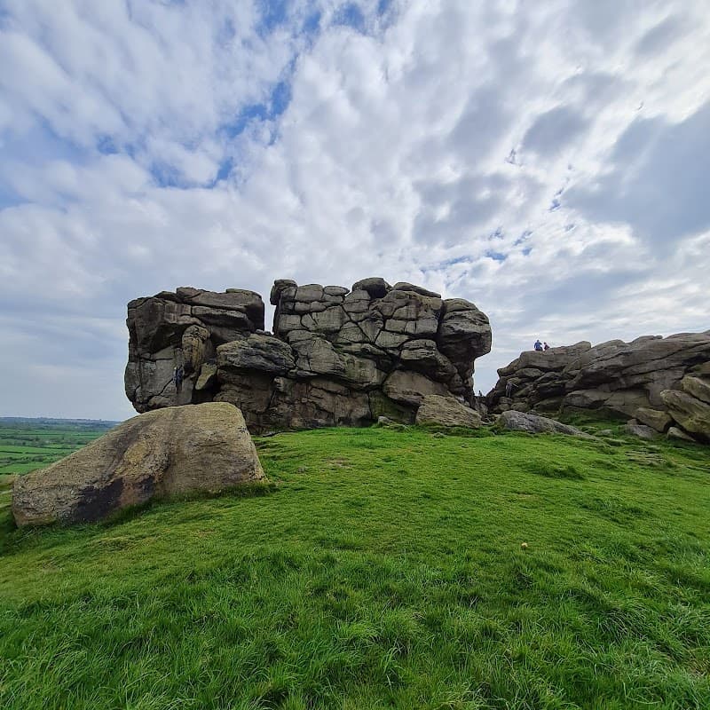 Large rocky outcrop on green grass with cloudy sky, featuring two people near the top of the crag.