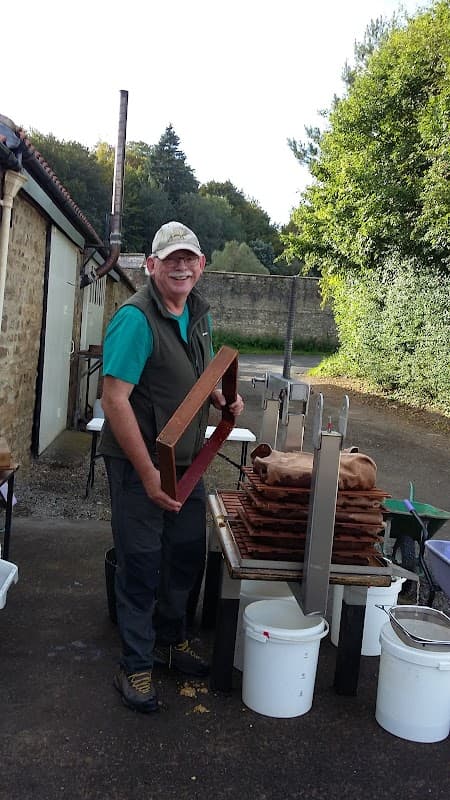 A smiling man holds a wooden frame near tables with trays and buckets in a farmyard setting surrounded by greenery.