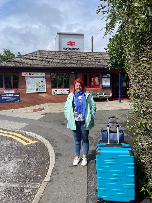 A woman with red hair stands near a blue suitcase outside Northallerton Station, with a sign and benches visible.