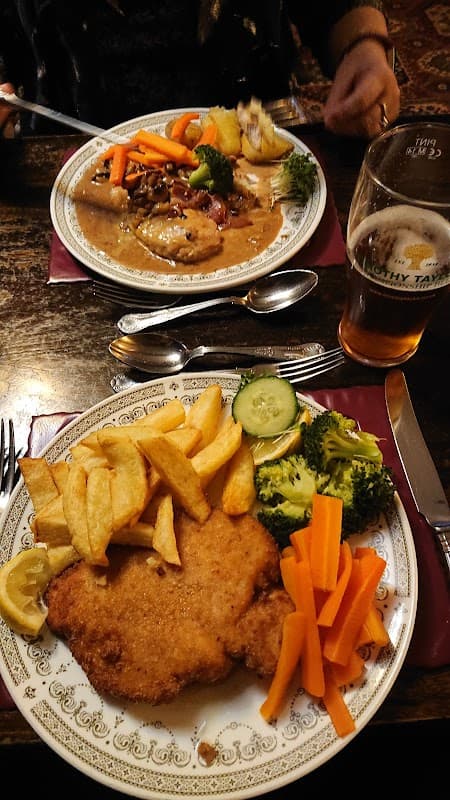 Two plates of hearty pub food with chips, vegetables, and a pint of beer on a wooden table in a cozy cafe setting.