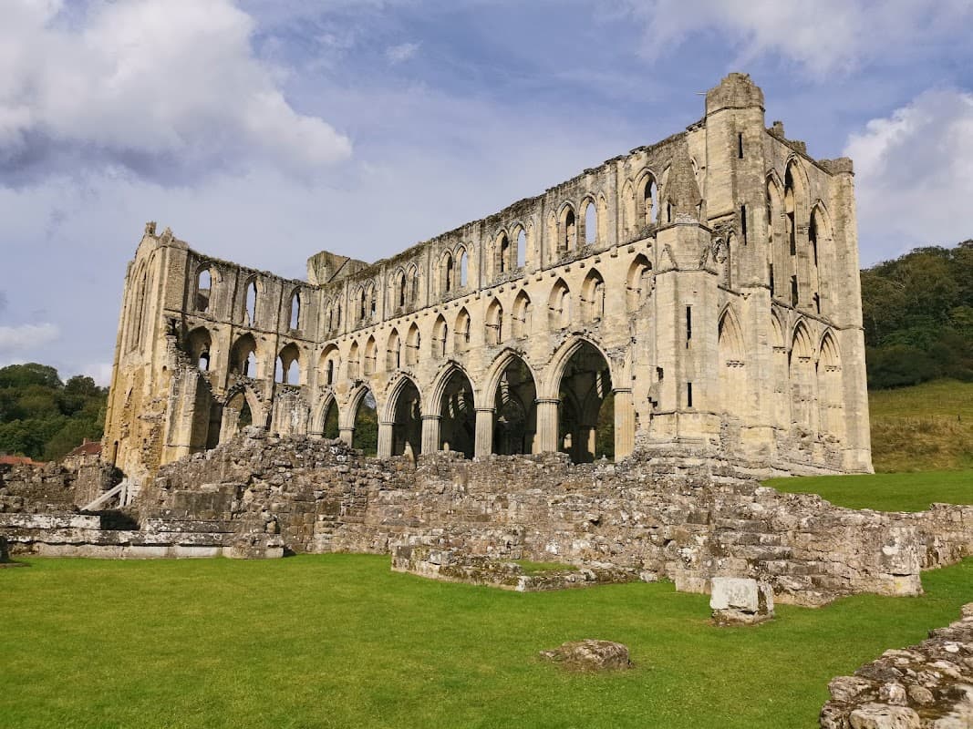 Ruins of Rievaulx Abbey with arched windows, stone walls, and a grassy foreground under a partly cloudy sky.