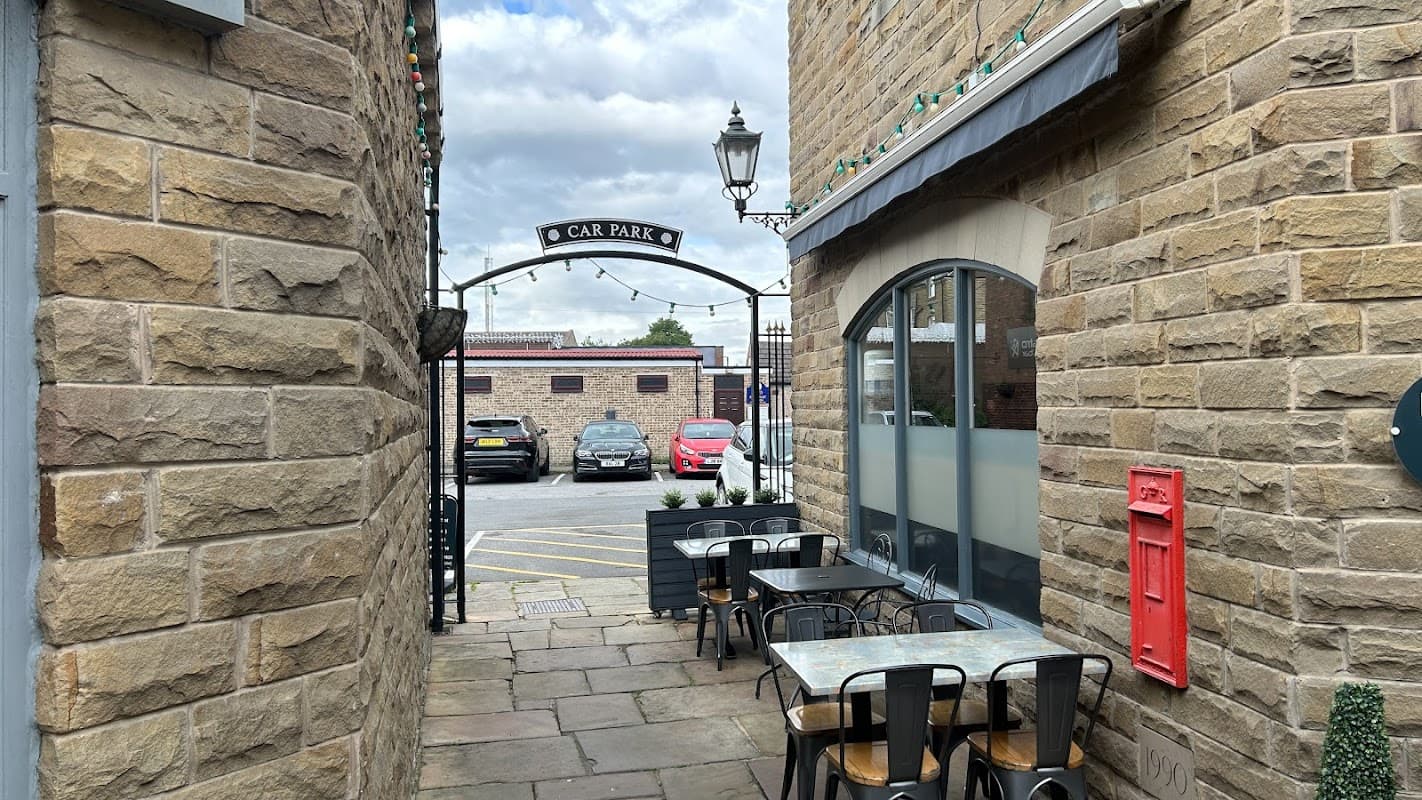 Stone walls frame a narrow passage leading to a car park, with tables, a red mailbox, and parked cars visible.