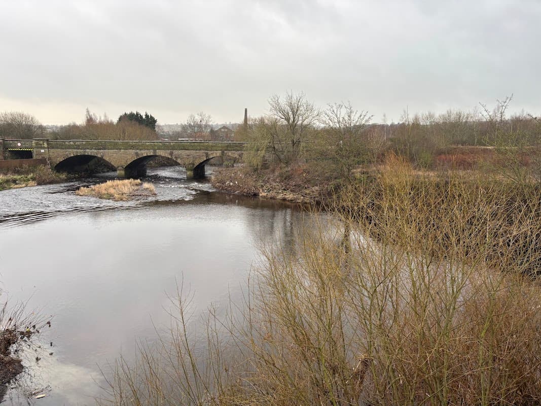 A tranquil river scene with a stone bridge, bare trees, and overcast skies in Ossett, Yorkshire.