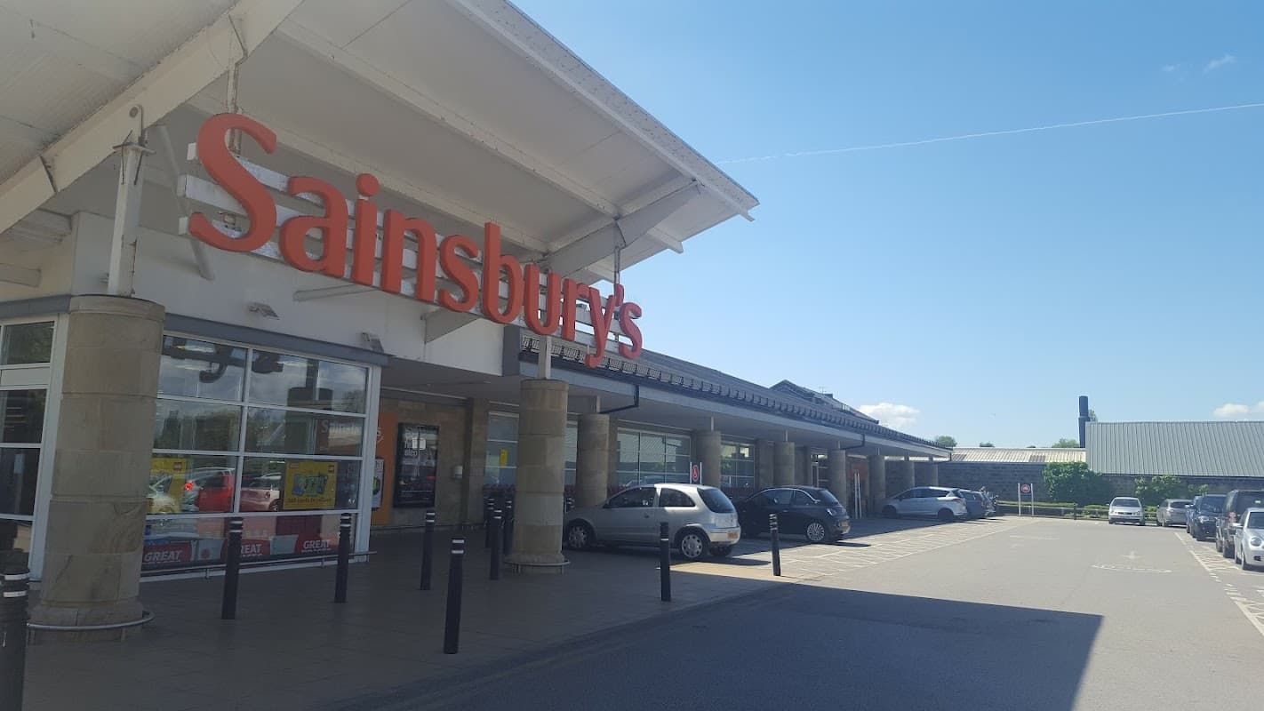 Sainsbury's storefront in Otley, Yorkshire, with cars parked outside under a clear blue sky.