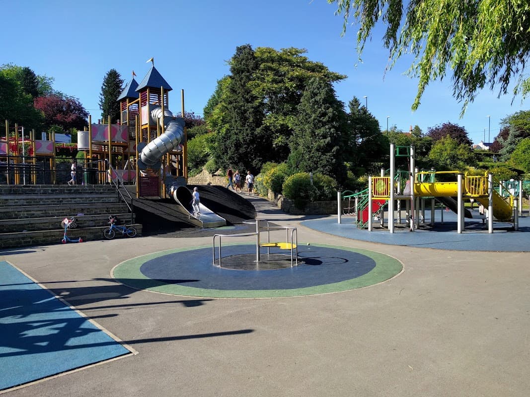 Playground featuring slides, climbing structures, and a spinning carousel, surrounded by greenery and a clear blue sky.