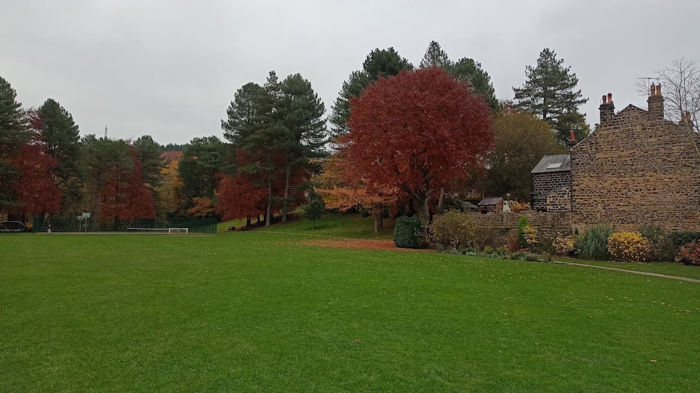 Lush green field surrounded by autumn trees, with vibrant red foliage and stone buildings in the background.