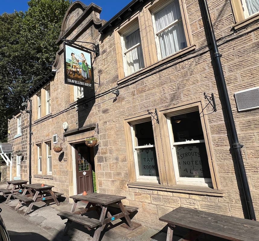 Stone building with large windows, wooden picnic tables outside, and a sign reading "Travellers Rest" above the entrance.
