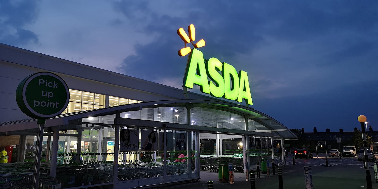 Brightly lit Asda store entrance with green signage and shopping trolleys outside, set against a twilight sky.