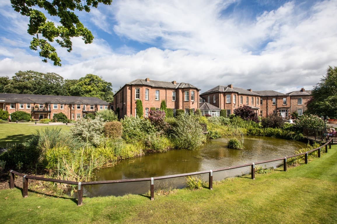 Retirement village with brick buildings, lush greenery, and a serene pond under a partly cloudy sky.
