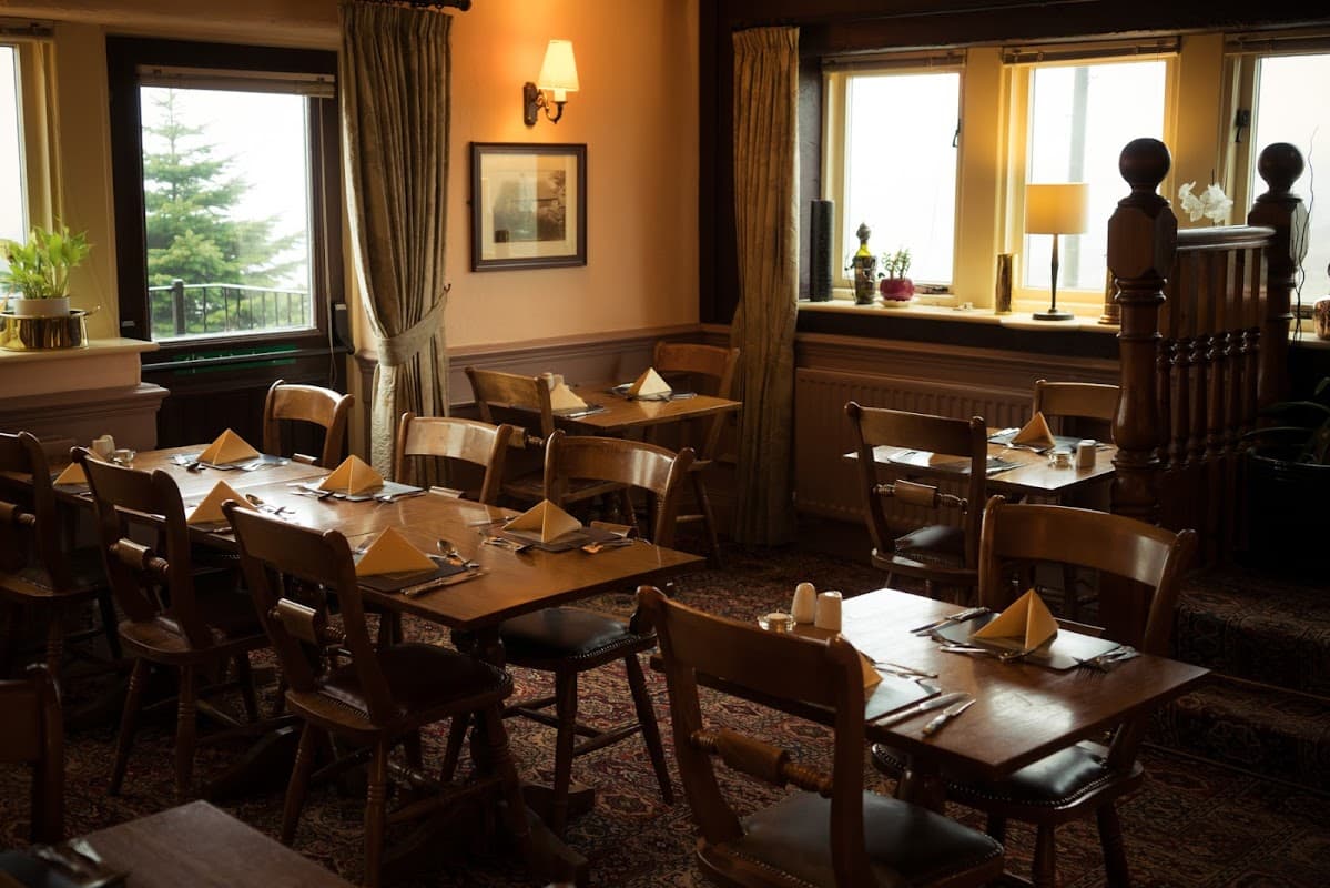 Cozy dining area with wooden tables, chairs, and neatly set napkins, surrounded by warm lighting and large windows.