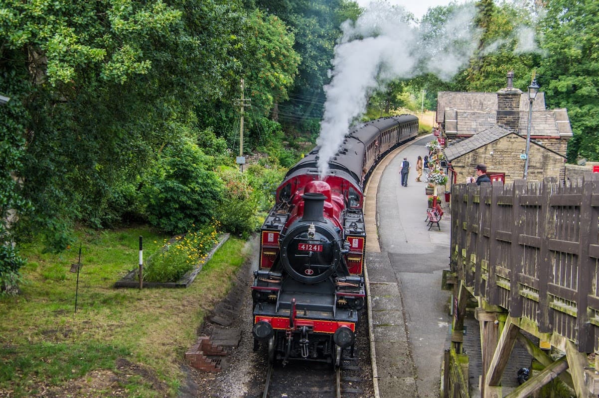 Steam train at Haworth station, surrounded by lush greenery and historic buildings, with passengers waiting nearby.