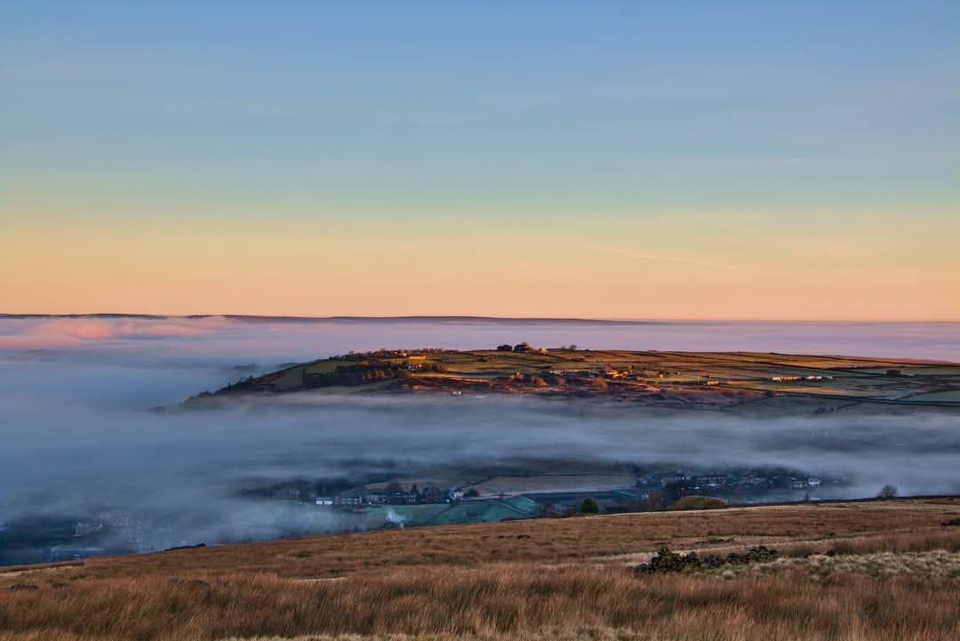 Rolling hills and valleys shrouded in mist, with a soft sunrise illuminating the landscape in Oxenhope, Yorkshire.