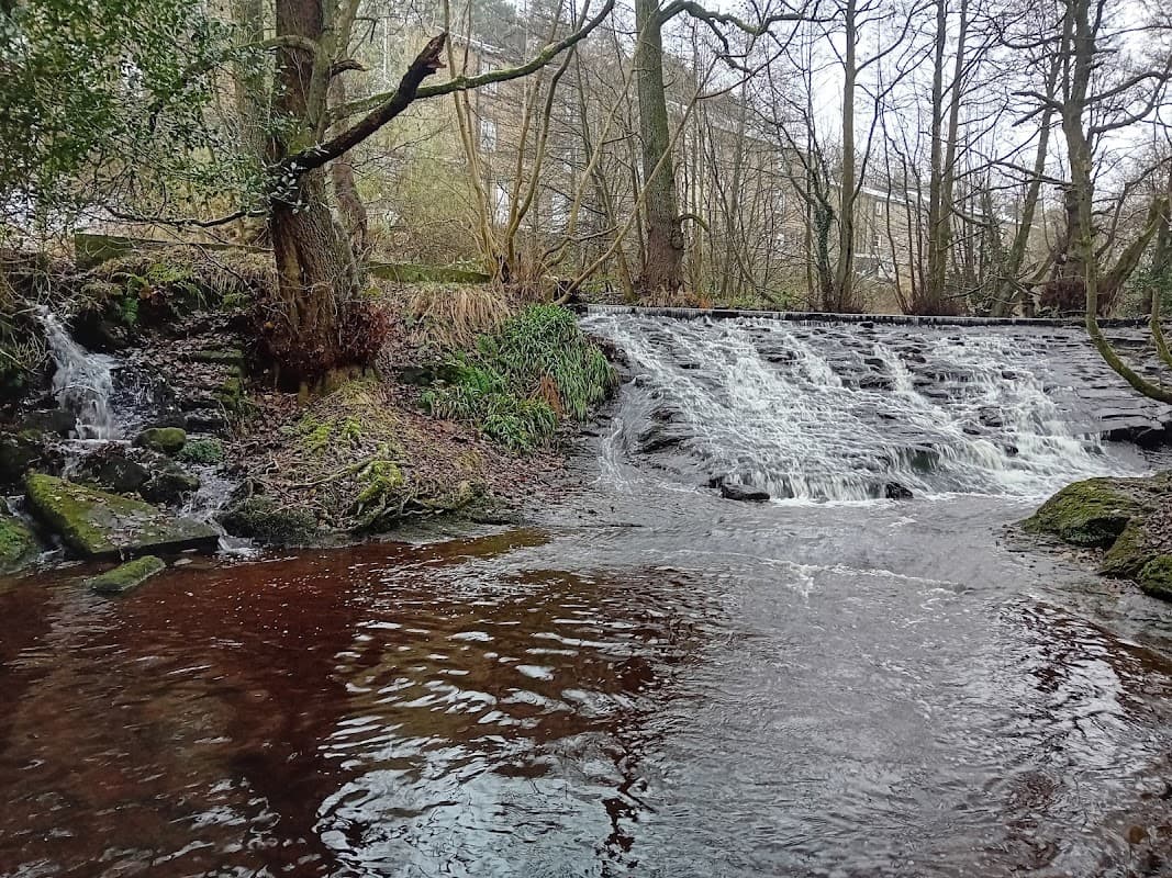 A serene waterfall cascades over rocks into a calm river, surrounded by trees and greenery in Oxenhope, Yorkshire.