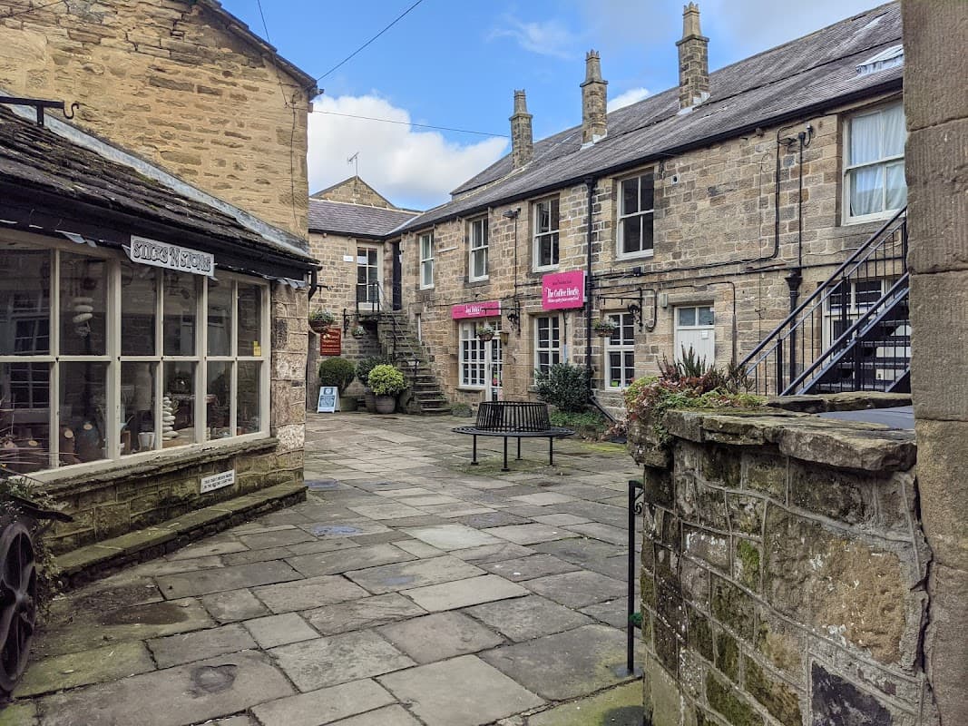 Quaint stone buildings with a courtyard, benches, and a staircase, featuring shops and a blue sky above.
