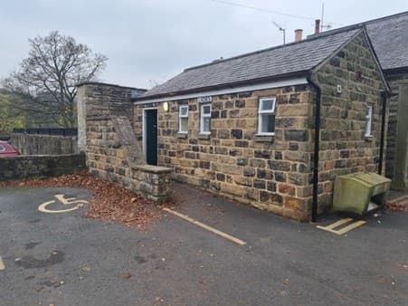 Stone building with a sloped roof, accessible parking space, and autumn leaves scattered on the ground.