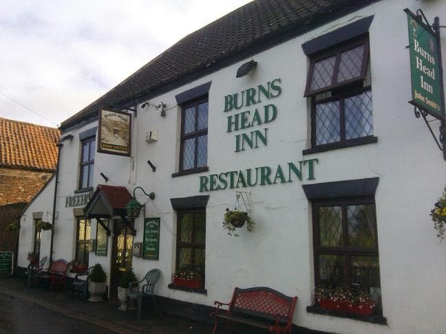 Traditional pub exterior with "Burns Head Inn" sign, green lettering, flower baskets, and benches outside.