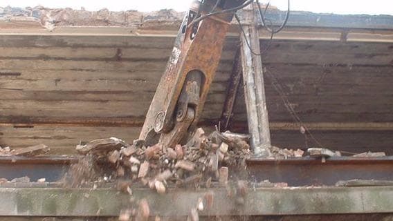 A demolition machine's claw dropping debris from a building's roof, with exposed wooden beams in the background.