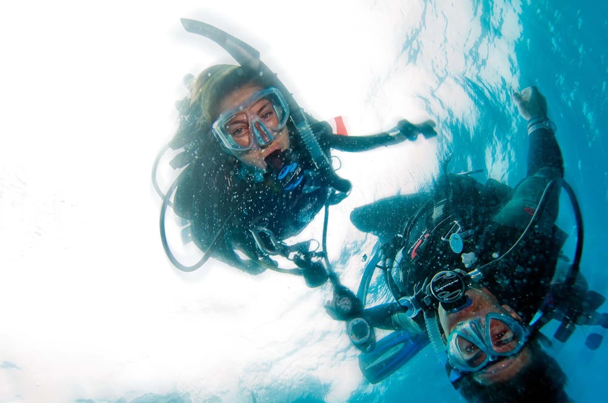 Two divers underwater, wearing scuba gear and masks, surrounded by bubbles and clear blue water.