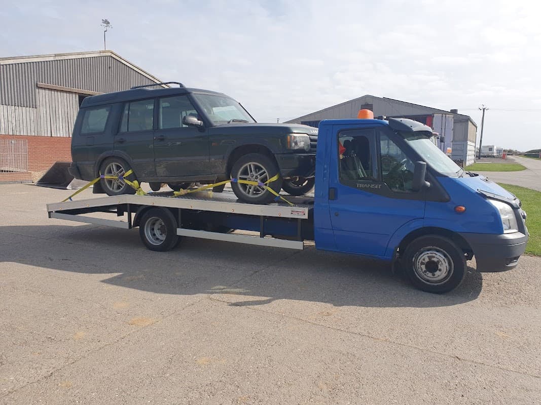 A blue tow truck carrying a green vehicle on a flatbed, with storage buildings in the background.