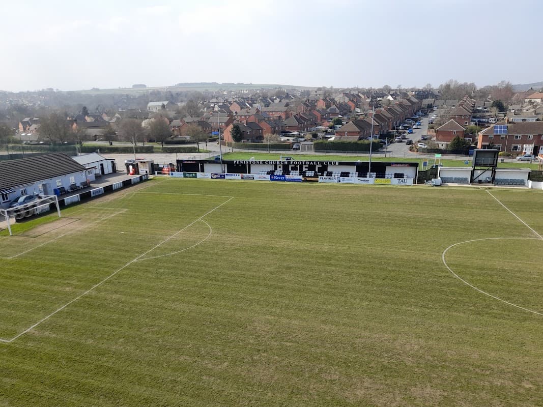 Green football pitch with stands, club signage, and residential area in the background under a clear sky.