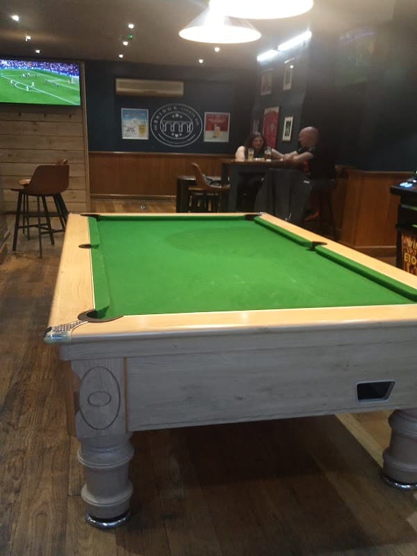 Pool table in a cozy pub setting with a football match on TV and patrons seated at a table in the background.