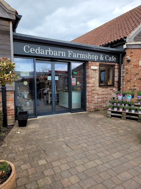 Cedar Barn Farm Shop & Cafe entrance with glass doors, brick walls, and potted plants in front.