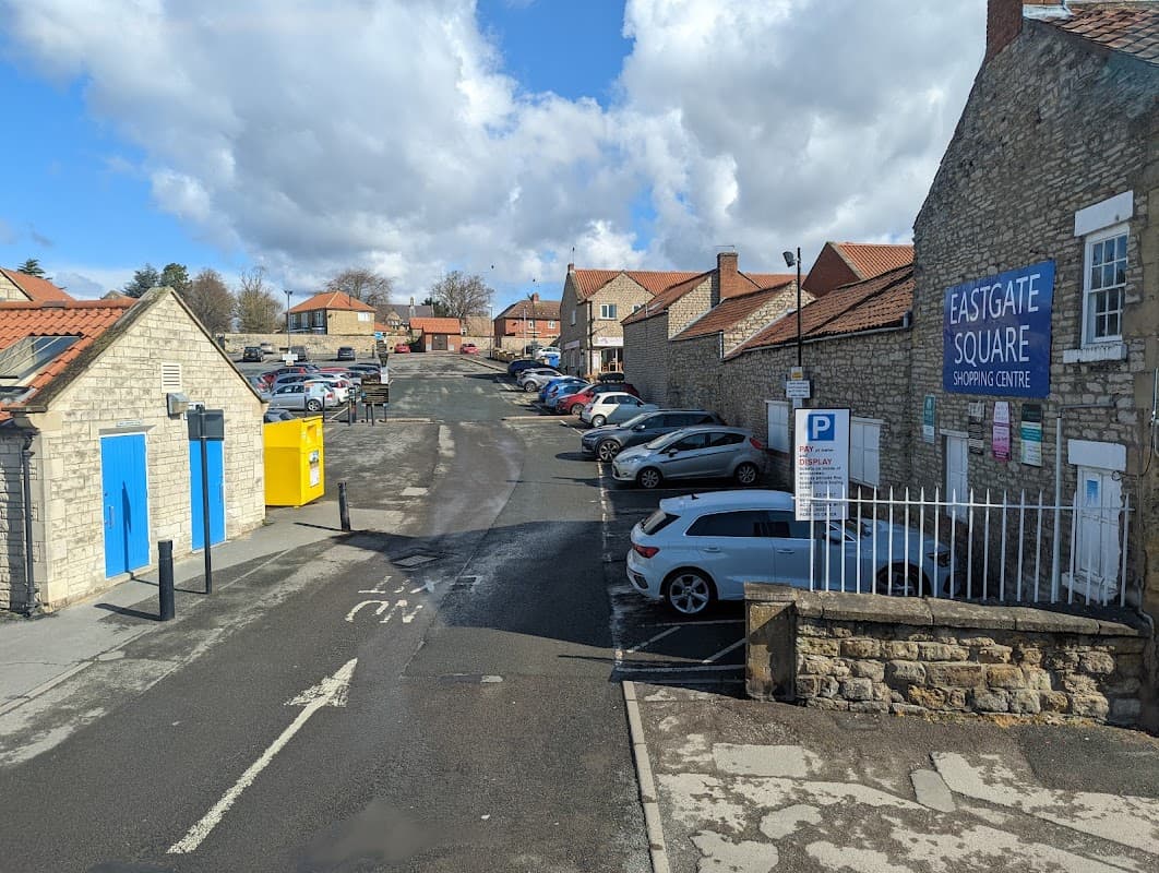 Eastgate Car Park with cars parked, stone buildings, blue sky, and clouds, near Eastgate Square Shopping Centre.