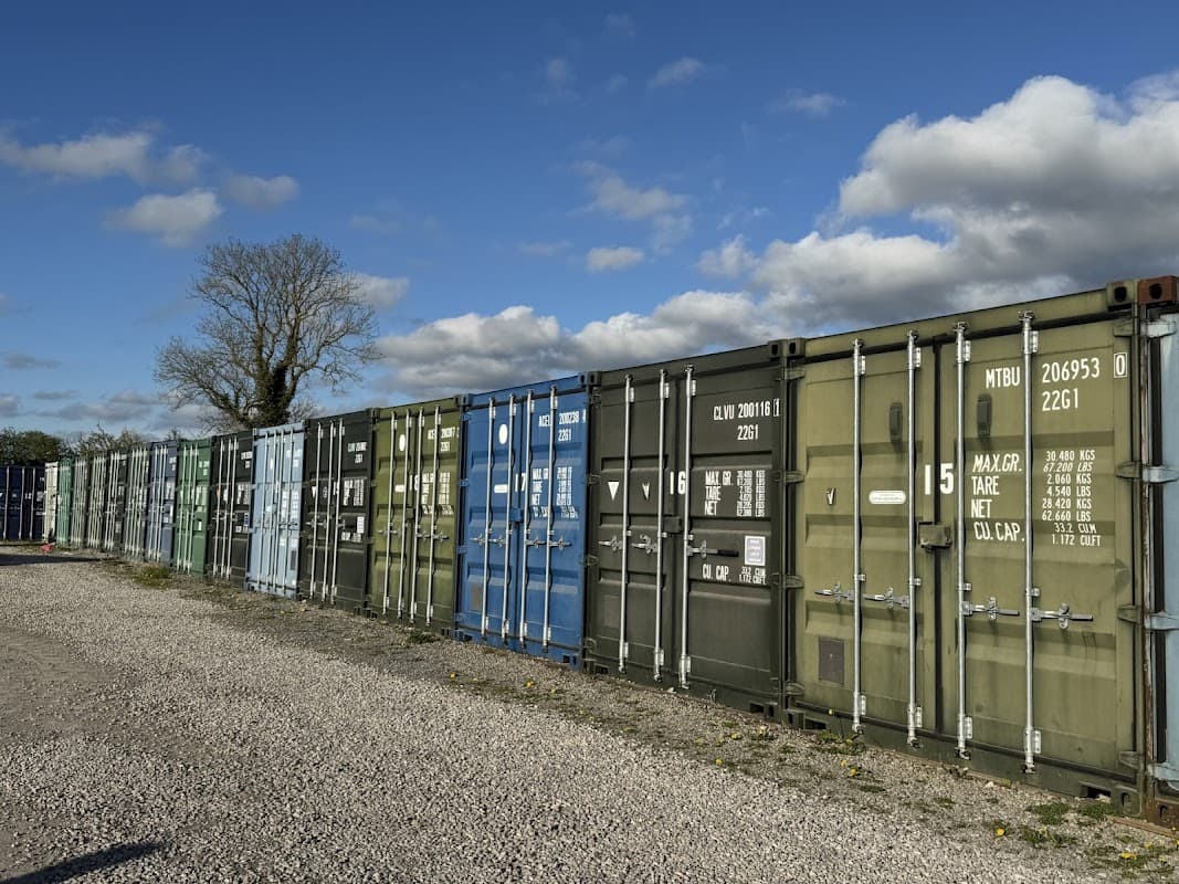 Colorful shipping containers lined up in a gravel yard under a partly cloudy sky, with a tree in the background.