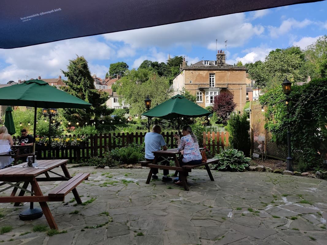 Two people sit at a picnic table under green umbrellas, surrounded by a garden and historic buildings in Plompton.