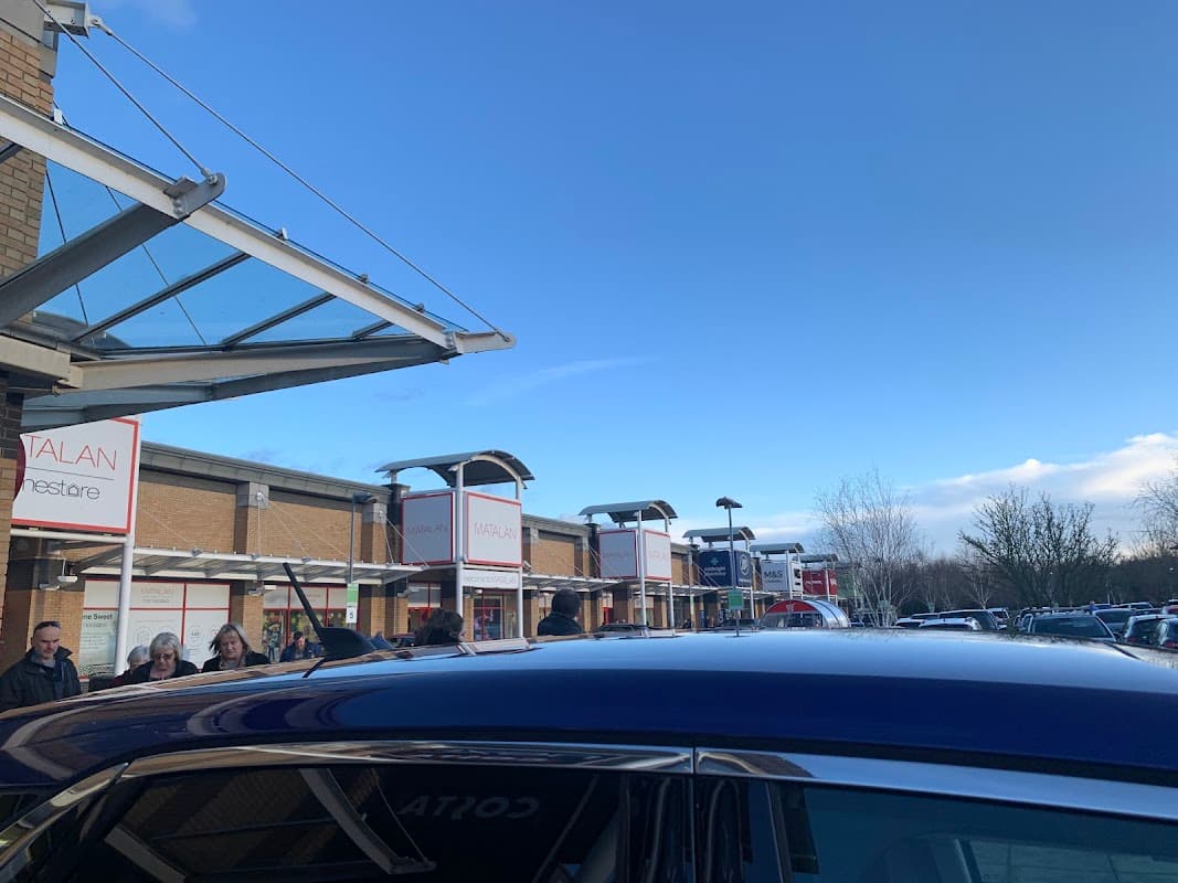 Shops and car park at St James Retail Park under a clear blue sky, with people walking and parked cars visible.