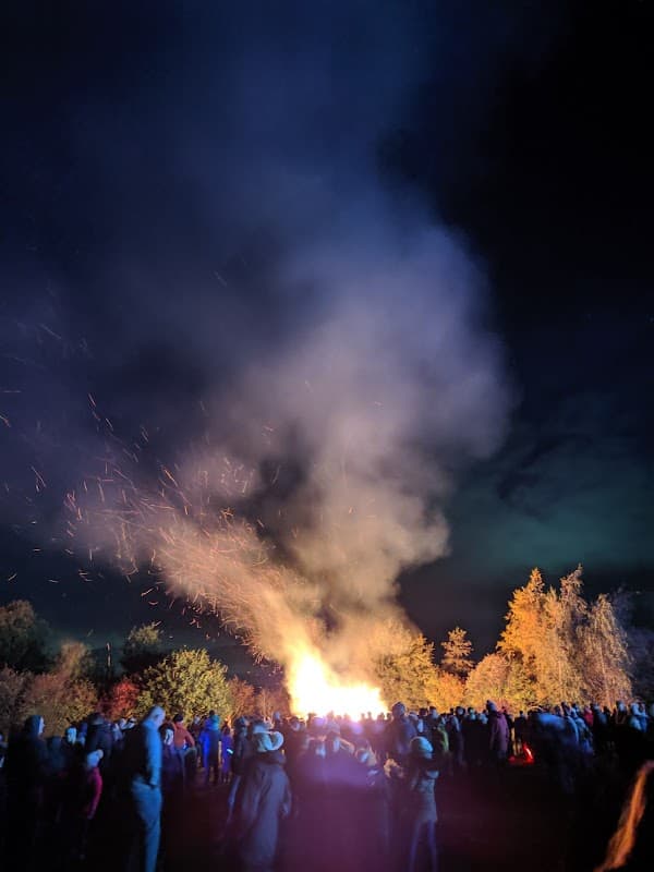 Crowd gathered around a large bonfire with smoke and sparks rising into the night sky, surrounded by trees.