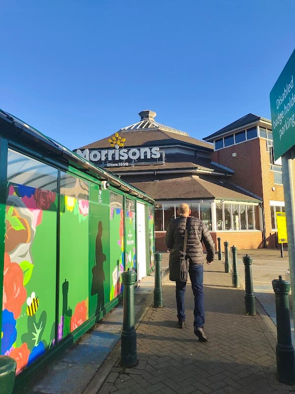 A person walks towards Morrison's Supermarket, with colorful floral murals and clear blue skies in the background.