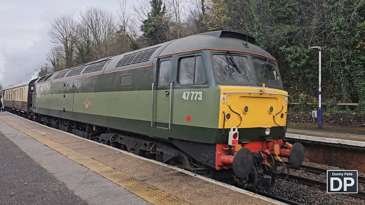 A green and yellow train engine at a station platform surrounded by trees and a gravel area.