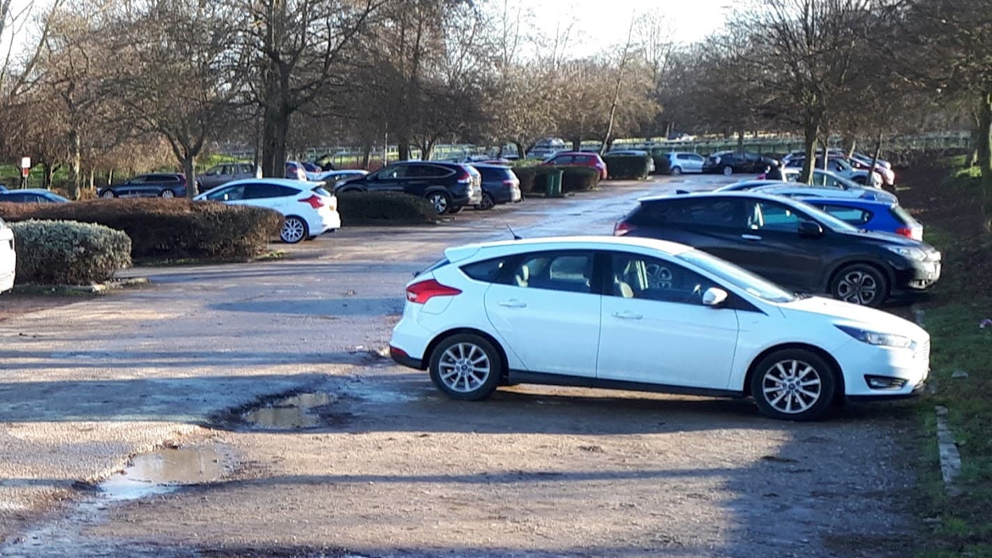 A busy car park with various parked cars, trees in the background, and a muddy patch on the ground.