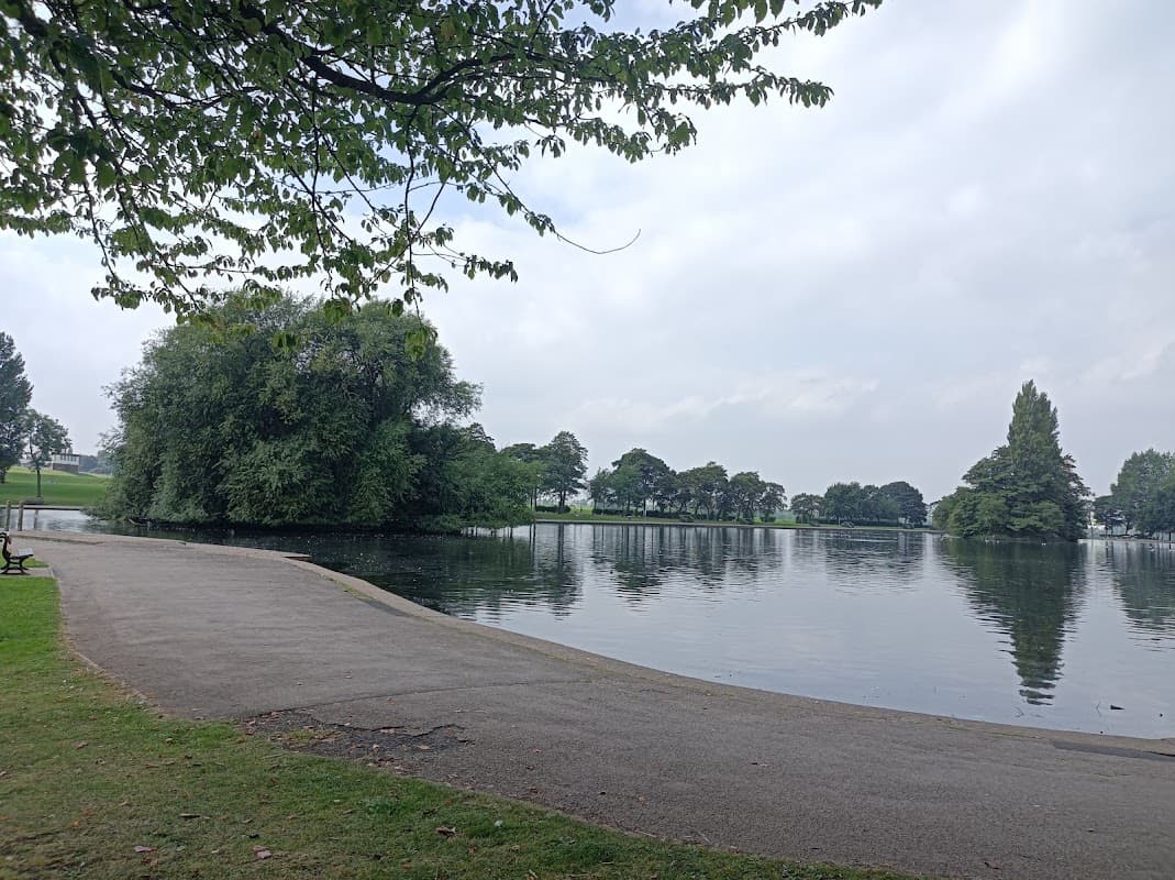 Serene park scene featuring a calm lake, lush greenery, and a pathway lined with trees under a cloudy sky.