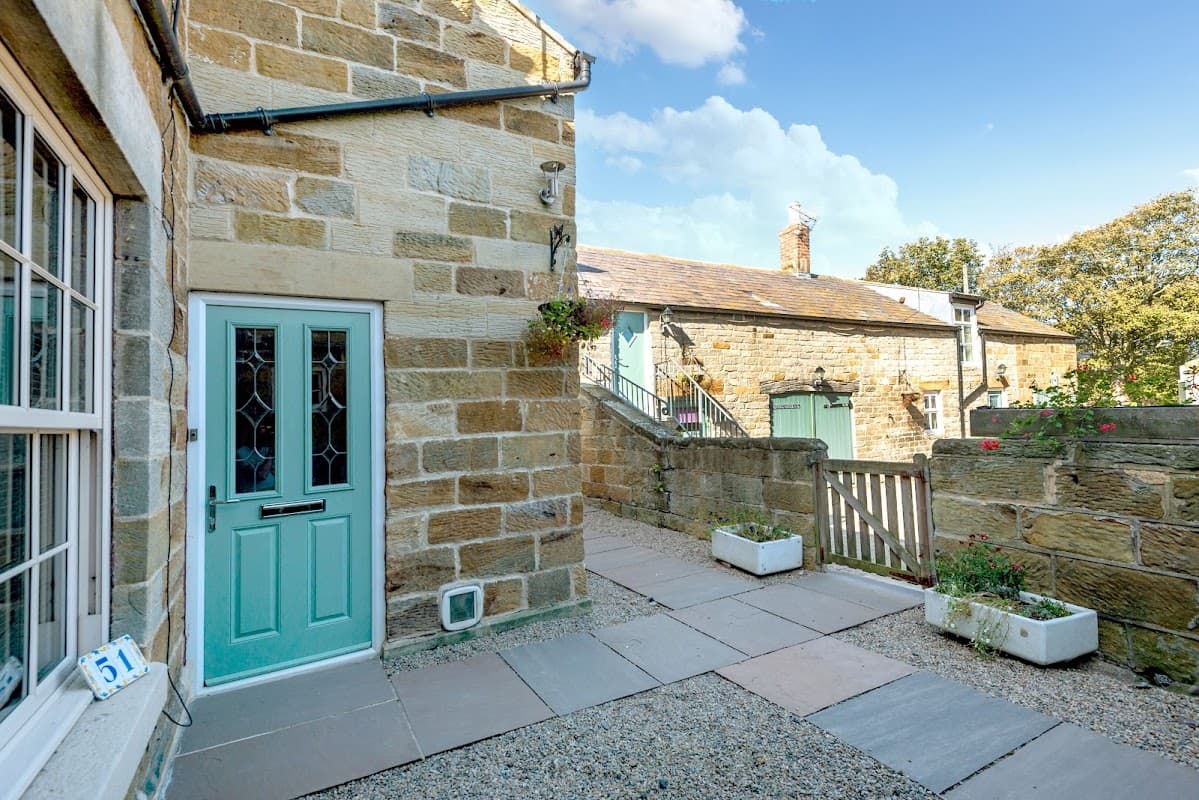 Charming stone cottage entrance with a teal door, pathway, and lush greenery in Port Mulgrave, Yorkshire.