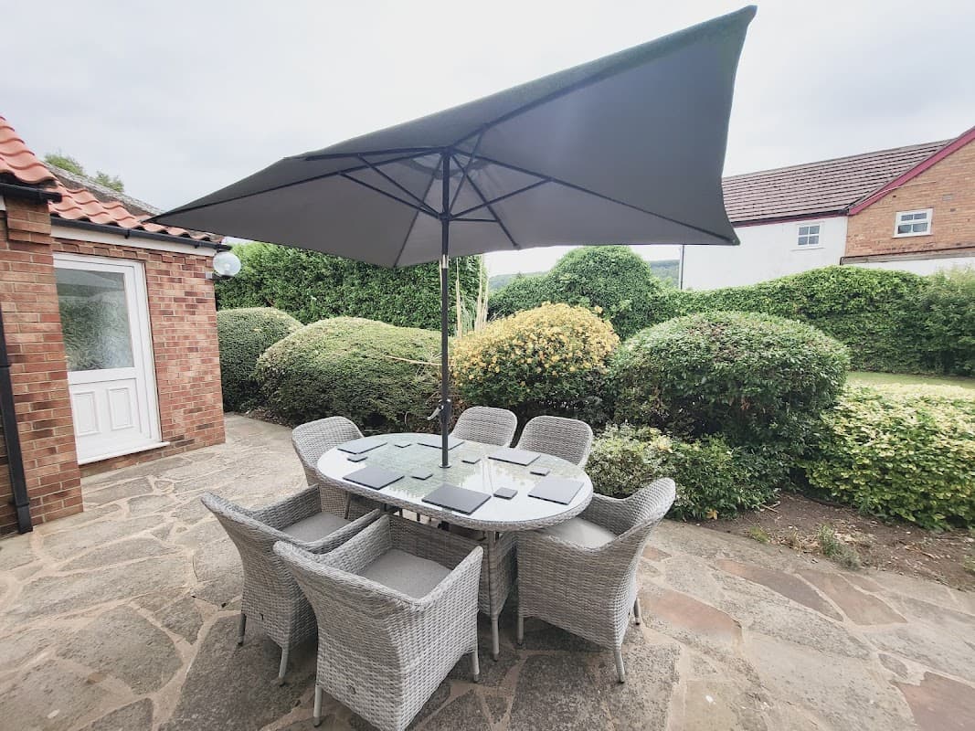 Outdoor seating area with a round table and eight wicker chairs under a large umbrella, surrounded by greenery.