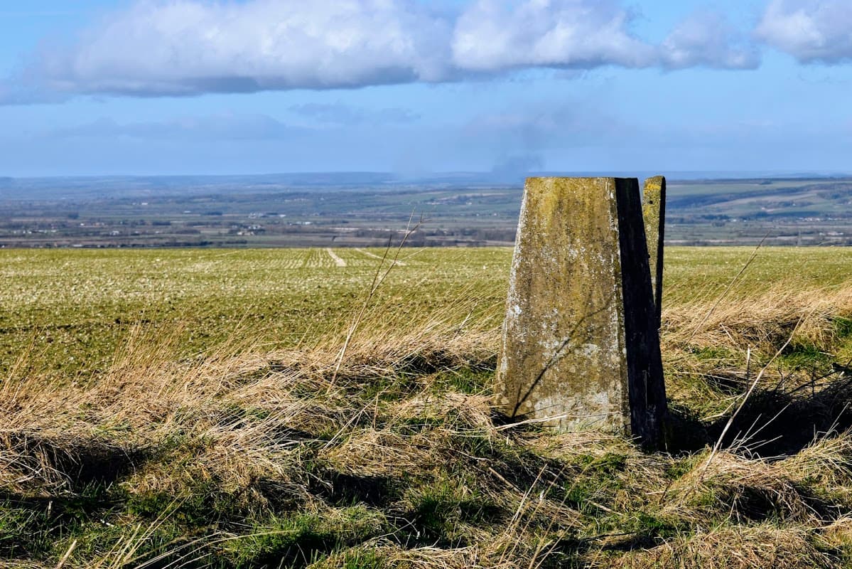 Trig point stands in a grassy field, overlooking a vast landscape with rolling hills under a partly cloudy sky.