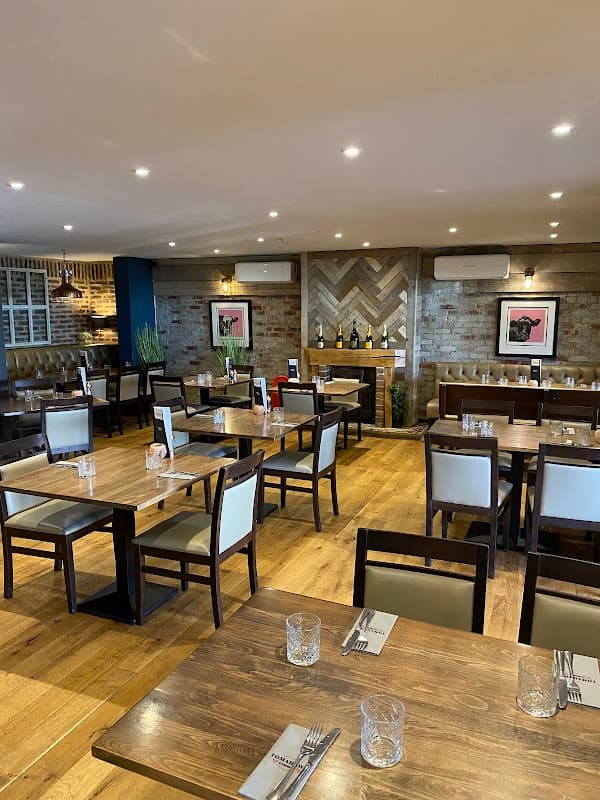Cozy restaurant interior with wooden tables, chairs, and a stylish bar area featuring wine bottles and artwork.