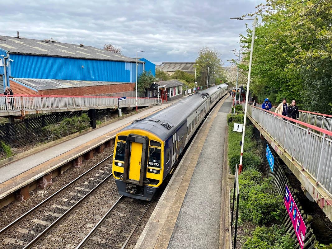 New Pudsey Station with a train approaching, platforms on either side, and people waiting. Green trees in the background.